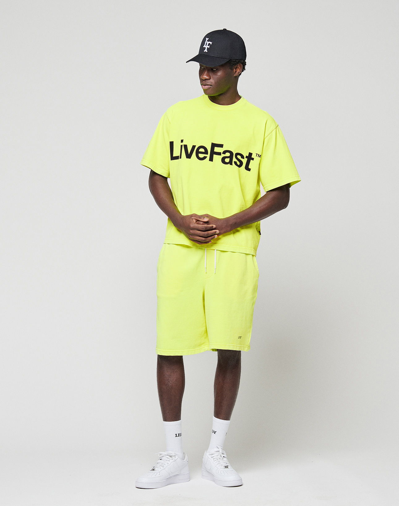 A person wears a neon yellow LIVE FAST DIE YOUNG Basic OS Livefast Tee with front print, matching shorts, black cap, white crew socks, and white sneakers against a plain light gray background. The oversize tee is 100% Baumwolle.
