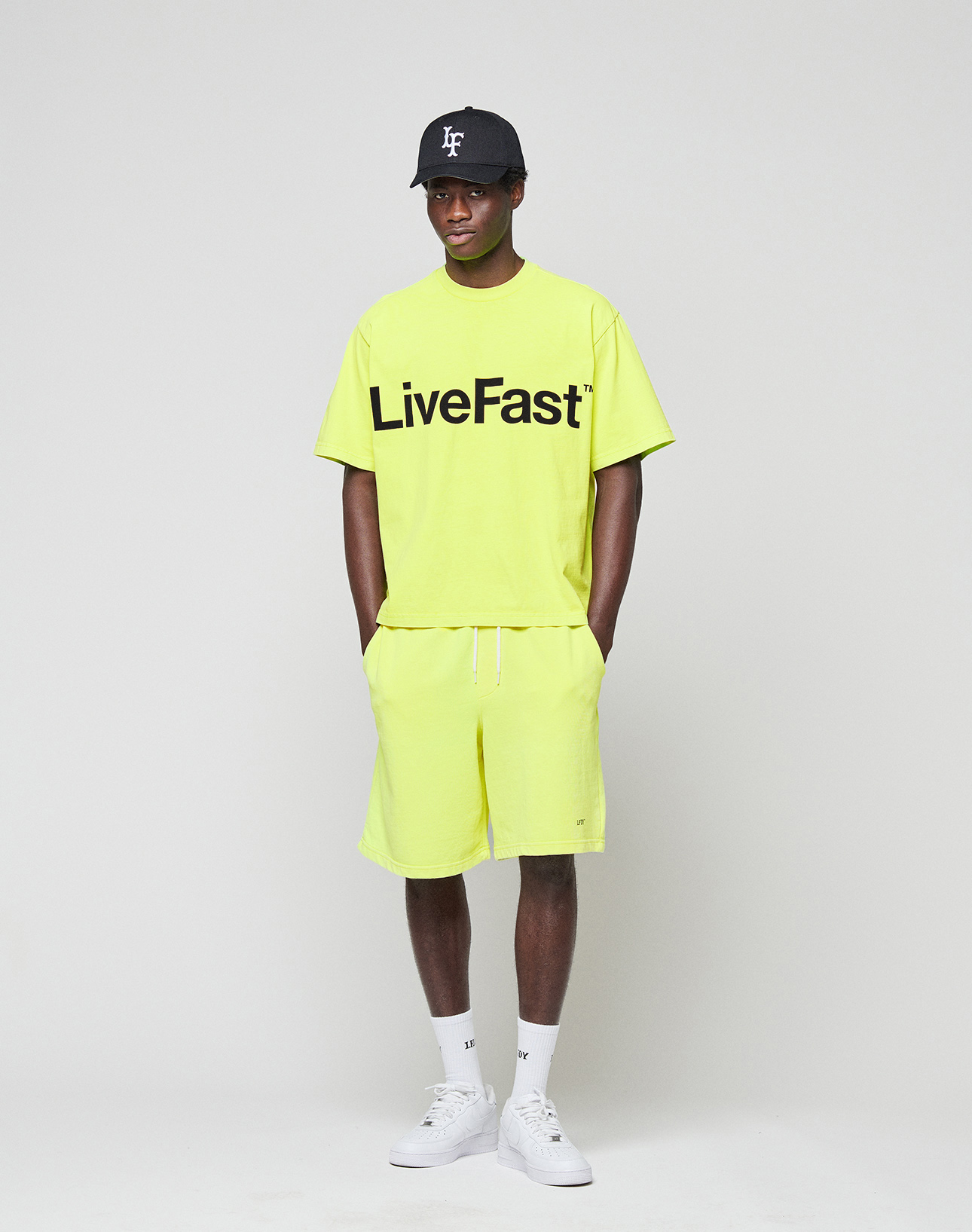 A person stands against a plain background, wearing a neon yellow Regular Fit LIVE FAST DIE YOUNG t-shirt with logo print, Basic Shorts with elastic waistband, white socks, white sneakers, and a black baseball cap. Their hands are in their pockets.