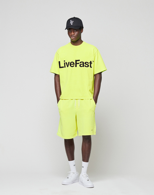 A person stands against a plain background, wearing a neon yellow Regular Fit LIVE FAST DIE YOUNG t-shirt with logo print, Basic Shorts with elastic waistband, white socks, white sneakers, and a black baseball cap. Their hands are in their pockets.