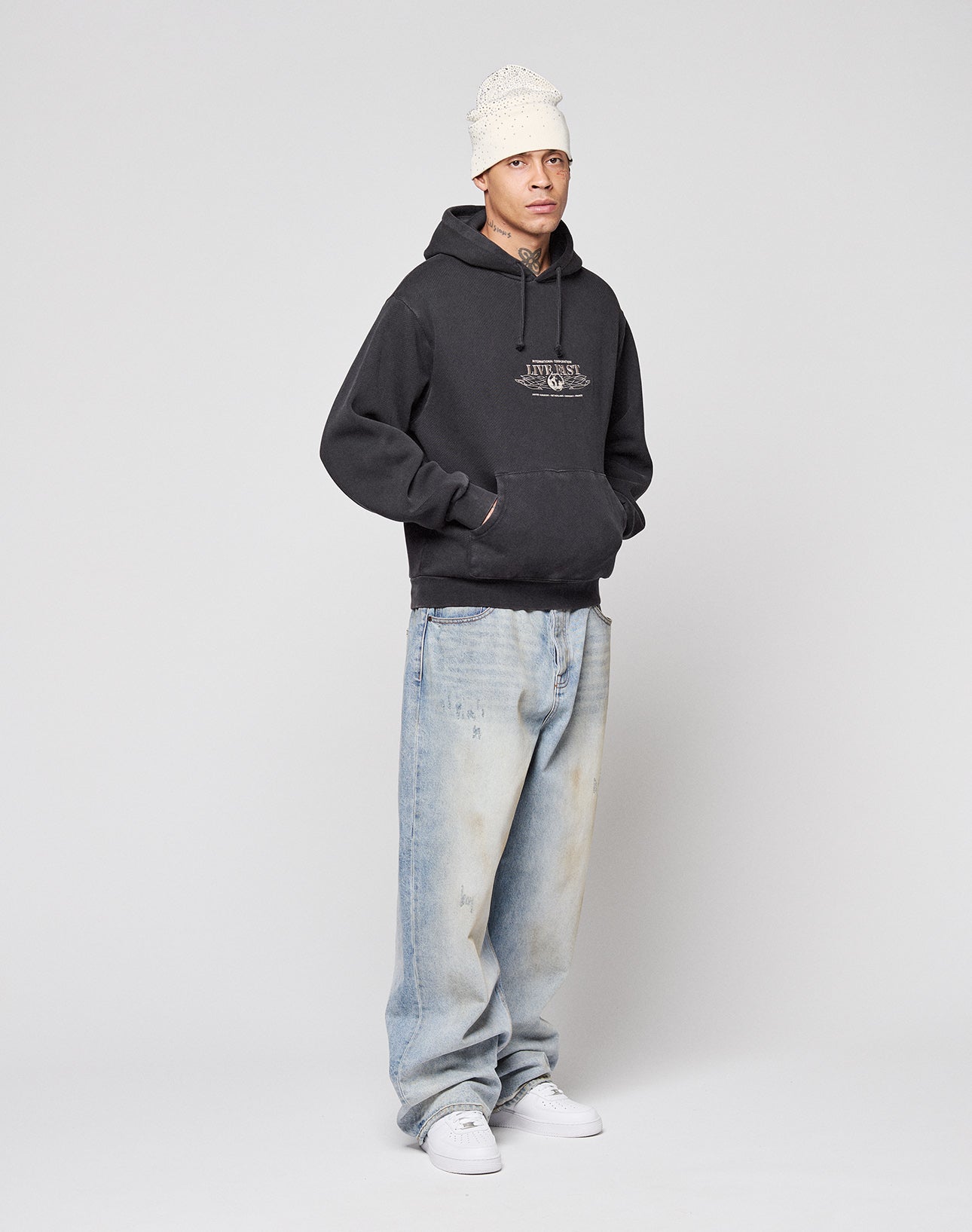 A person wears a white knit beanie, LIVE FAST DIE YOUNG's Live Fast Corporation Hooded with print artwork, loose-fitting light blue jeans in Vintage Waschung, and white sneakers, standing hands-in-pocket against a plain light background.