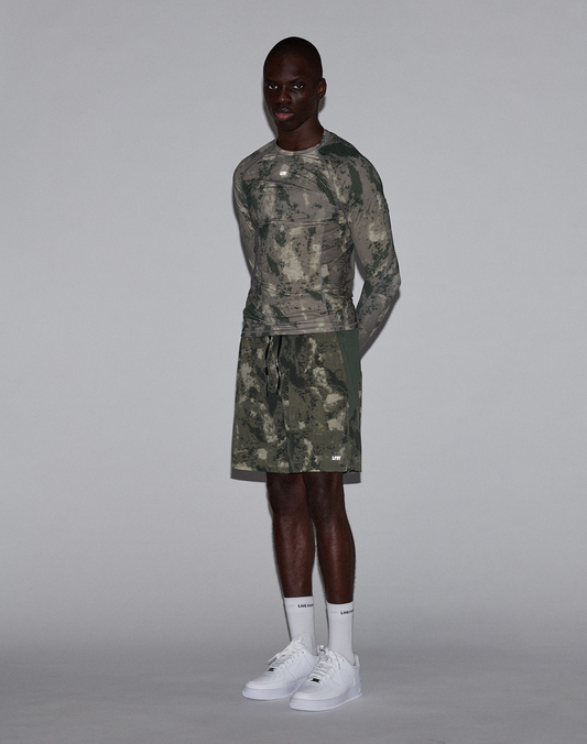 A man stands against a plain gray backdrop wearing LIVE FAST DIE YOUNG LFDY Performance Running Shorts, a green and gray camo long-sleeve shirt with reflective logo print, white socks, and white sneakers. He faces forward with arms relaxed.