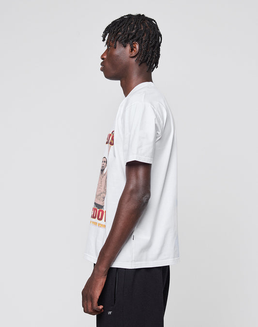 A young man with short dreadlocks wears the Abus Magomedov Tee by LIVE FAST DIE YOUNG, a white T-shirt with front and back print in 100% Baumwolle, paired with black shorts, standing in profile against a plain light gray background.
