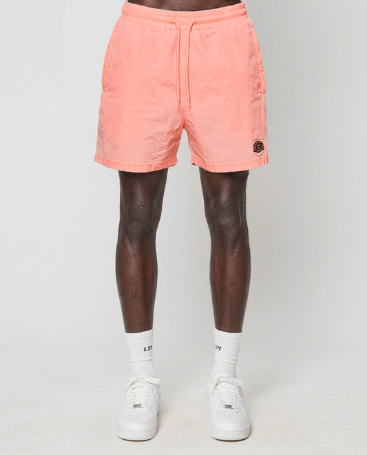 A person models LIVE FAST DIE YOUNG Live Fast Swim Shorts in light pink peach, paired with white crew socks and sneakers against a plain background. Only the lower half of their body is visible.