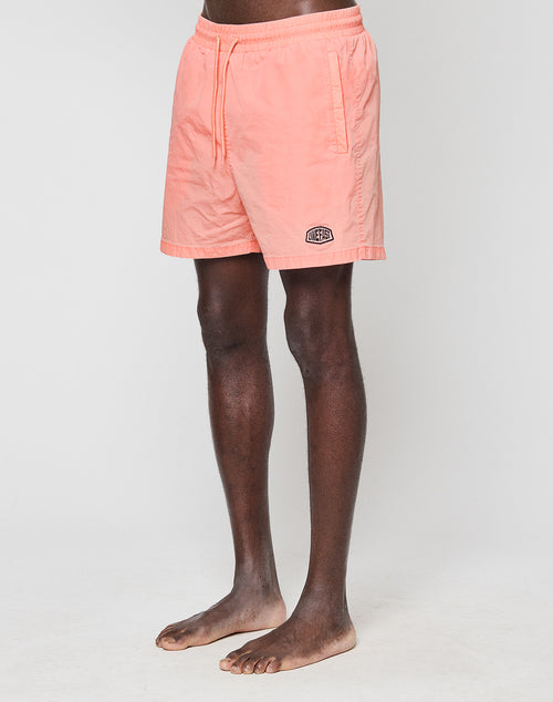 A person stands barefoot against a white background wearing LIVE FAST DIE YOUNG’s light pink Live Fast Swim Shorts, featuring an elastic waistband, side pockets, and a small logo above the left hem.