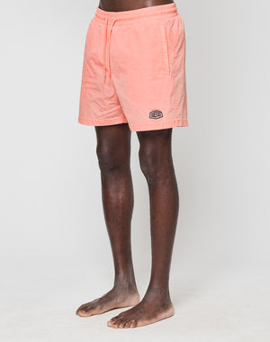 A person stands barefoot against a white background wearing LIVE FAST DIE YOUNG’s light pink Live Fast Swim Shorts, featuring an elastic waistband, side pockets, and a small logo above the left hem.