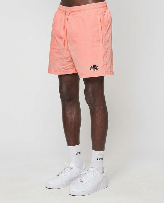 A person wearing LIVE FAST DIE YOUNG Live Fast Swim Shorts in tinted peach wash, white crew socks with LPSD text, and white sneakers stands against a plain white background.