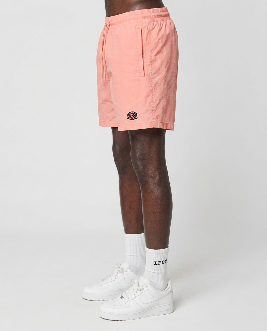 A person stands sideways against a plain white background, wearing LIVE FAST DIE YOUNG Live Fast Swim Shorts in Tinted Peach Wash with elastic waistband, white socks with LFDY text, and white sneakers.