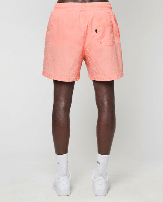 A person stands facing away, wearing LIVE FAST DIE YOUNG Live Fast Swim Shorts with an elastic waistband, white crew socks, and white sneakers against a plain light background. The shorts have a back pocket on the right side.