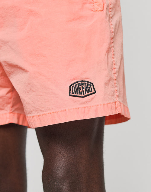 Close-up of LIVE FAST DIE YOUNG’s Live Fast Swim Shorts in light pink, featuring an embroidered LIVE FAST patch near the hem. Made from polyamide with an elastic waistband, these regular-fit shorts are set against a plain backdrop.