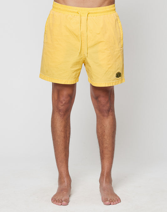 A person stands barefoot against a plain background, wearing LIVE FAST DIE YOUNGs Live Fast Swim Shorts—bright yellow, regular fit, wide elastic waistband, side pockets, and a small black circular logo on the left leg.