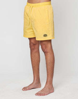 A person stands barefoot against a plain background, wearing LIVE FAST DIE YOUNG’s Live Fast Swim Shorts in tinted yellow with a wide elastic waistband, drawstring, and a small patch logo on the left leg. Their upper body and face are not visible.