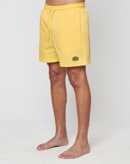 A person stands barefoot against a plain background, wearing LIVE FAST DIE YOUNG’s Live Fast Swim Shorts in tinted yellow with a wide elastic waistband, drawstring, and a small patch logo on the left leg. Their upper body and face are not visible.