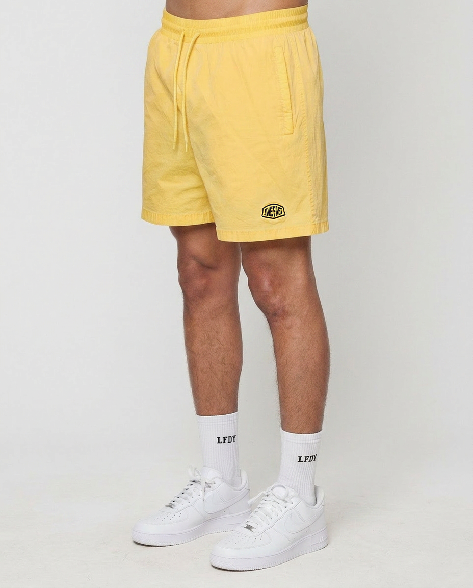 Only the lower body is visible as a person stands against a white background, wearing LIVE FAST DIE YOUNG Live Fast Swim Shorts in tinted yellow wash, white LFDY crew socks, and white sneakers.