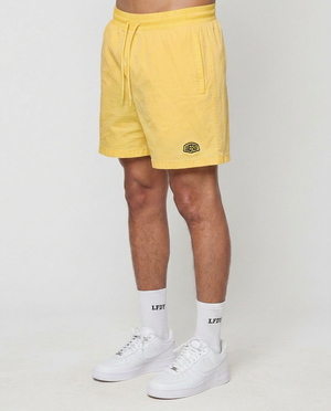 Only the lower body is visible as a person stands against a white background, wearing LIVE FAST DIE YOUNG Live Fast Swim Shorts in tinted yellow wash, white LFDY crew socks, and white sneakers.