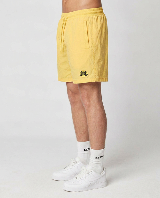 A person in profile wears LIVE FAST DIE YOUNG Live Fast Swim Shorts—regular fit, tinted yellow wash with an elastic waistband, pockets, and a round patch on the left leg—plus white LPDY socks and white sneakers against a plain light background.