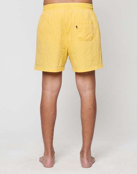 A person stands barefoot facing away from the camera, wearing LIVE FAST DIE YOUNG Live Fast Swim Shorts in a tinted yellow wash with a breiter Elastikbund and single back pocket, set against a plain, light background.