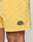 Close-up of a person wearing Live Fast Swim Shorts by LIVE FAST DIE YOUNG in Tinted Yellow Wash. These Regular Fit shorts feature a LIVE FAST patch near the hem and reveal part of the wearer’s thigh.