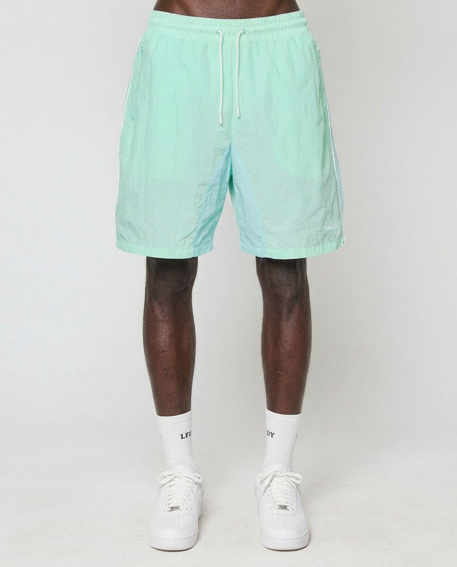 A person models LIVE FAST DIE YOUNG Beach Shorts in light teal with a drawstring and elastikbund, paired with white crew socks and white sneakers, standing against a plain white background. The image is cropped just below the waist.
