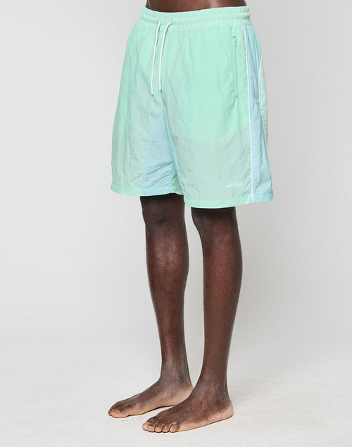 A person stands barefoot against a plain background, wearing LIVE FAST DIE YOUNG Beach Shorts in light green and pastel blue with elastic waistband, white drawstring, and side zip pockets. The upper body is not visible.