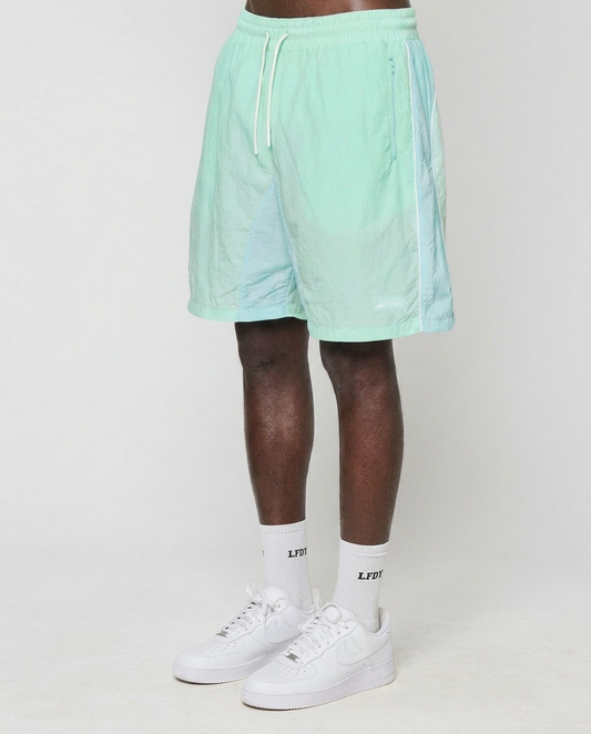 A person stands against a plain white background, wearing LIVE FAST DIE YOUNG Beach Shorts in mint green, white crew socks with LFDT branding, and white Nike sneakers. Only the lower half of their body is visible. | model