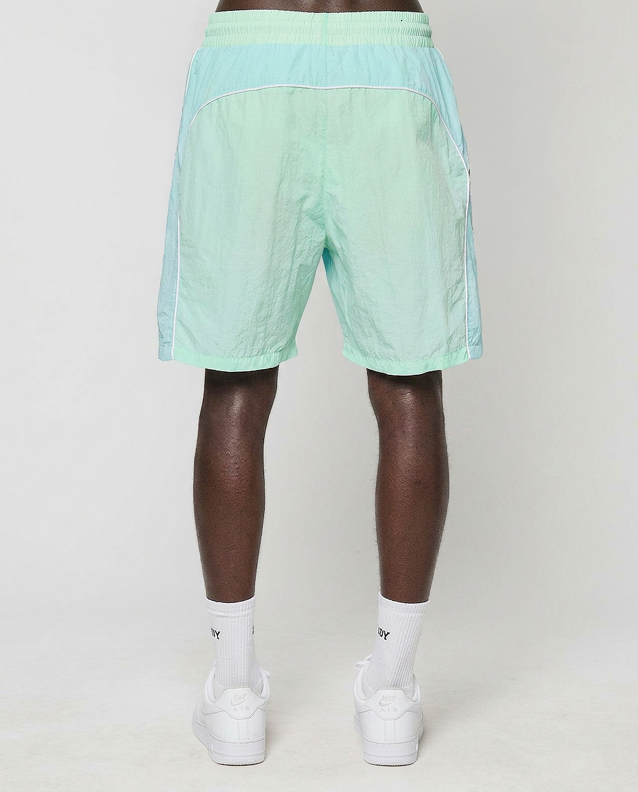 A person faces away from the camera wearing LIVE FAST DIE YOUNG Beach Shorts in light green with white trim and elastikbund, paired with white socks and sneakers, set against a plain light background.