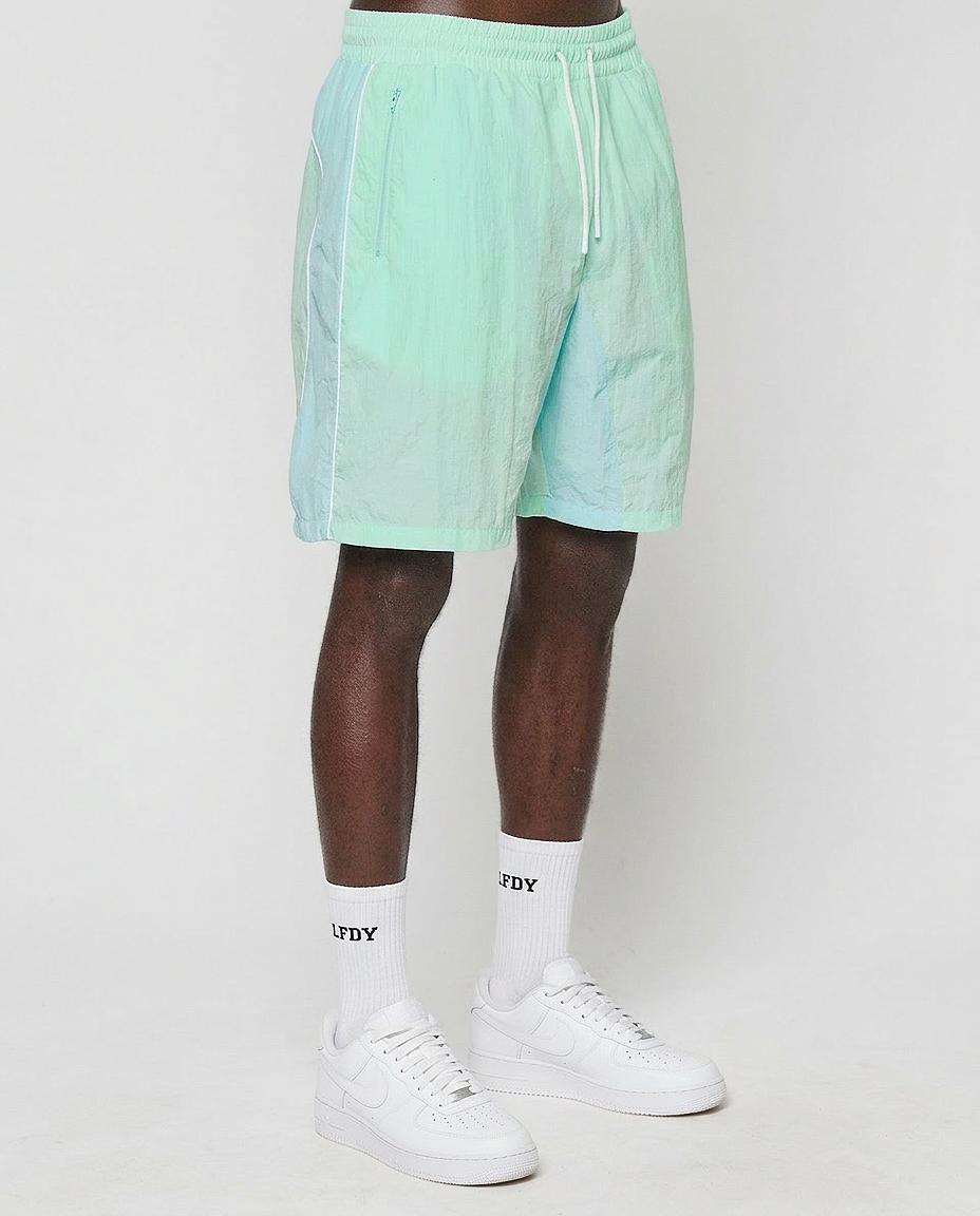 A person wears LIVE FAST DIE YOUNG Beach Shorts in pastel mint green with an elastic waistband, white crew socks featuring LFDY lettering, and white sneakers. The image, cropped above the waist, shows them standing against a plain white background.