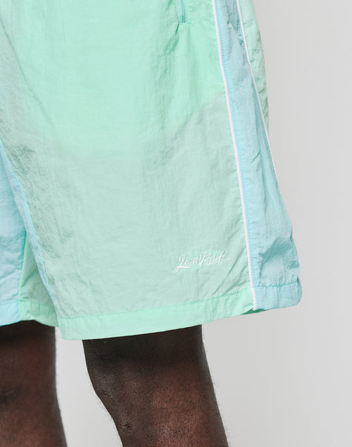 A close-up shows the lower part of LIVE FAST DIE YOUNG Beach Shorts in mint green and light blue colorblock with white piping, an elastic waistband, and “Live Fast” embroidered near the hem. Only the upper legs and shorts are visible.