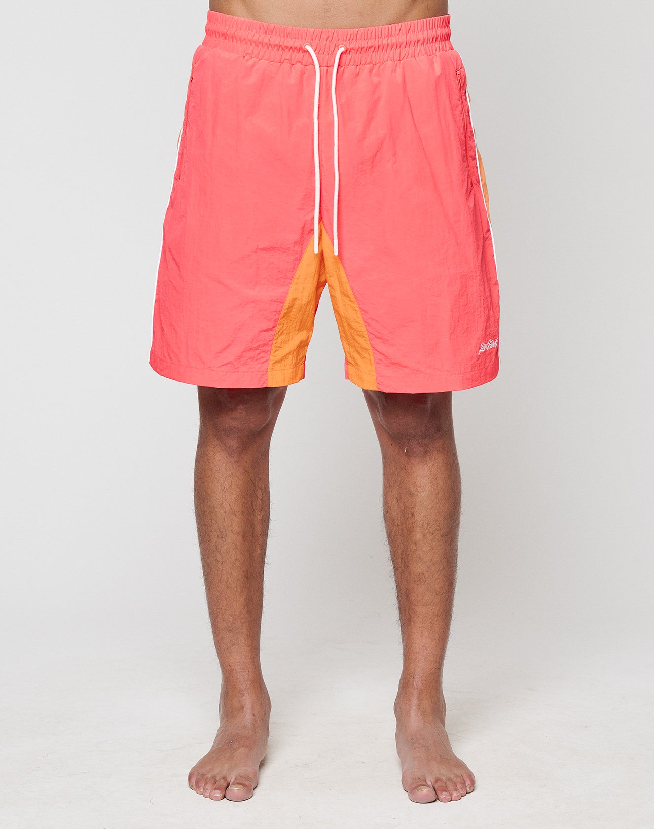 A barefoot person models LIVE FAST DIE YOUNG Beach Shorts in coral and orange colorblock with Breiter Elastikbund and white drawstrings, photographed from the waist down against a plain light background.