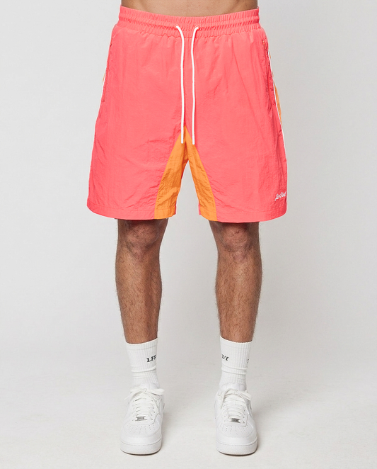 A person wears LIVE FAST DIE YOUNG Beach Shorts in bright coral and orange with Elastikbund and seitliche Eingrifftaschen, white crew socks, and white sneakers, standing against a plain light background. The shorts have a Regular Fit for comfort.