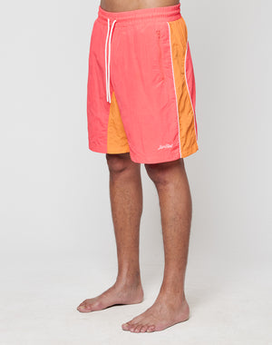 A person stands barefoot against a plain light background, wearing LIVE FAST DIE YOUNG Beach Shorts in coral and orange with side panels, Regular Fit, wide elastic waistband, drawstring, and a small embroidered logo on the left leg.