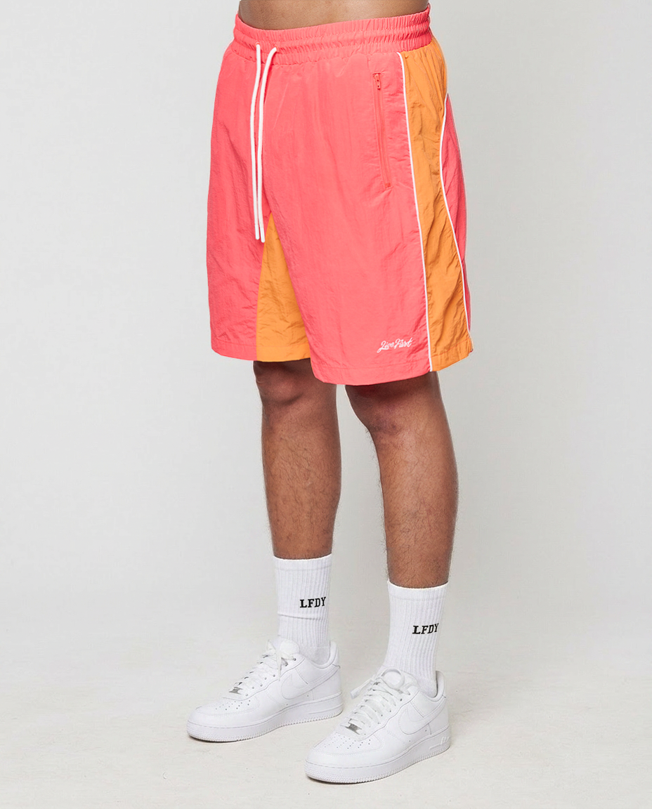 A person wears LIVE FAST DIE YOUNG Beach Shorts in orange and pink with side pockets and elastic waistband, white crew socks with LFDY print, and white sneakers, standing against a plain light background.