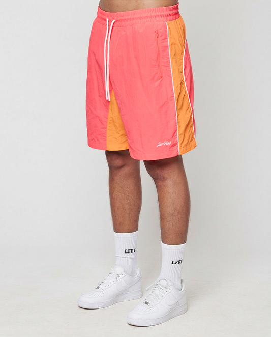 A person wears LIVE FAST DIE YOUNG Beach Shorts in orange and pink with side pockets and elastic waistband, white crew socks with LFDY print, and white sneakers, standing against a plain light background. | model