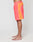 A person stands barefoot in profile against a white background, wearing LIVE FAST DIE YOUNG Beach Shorts in coral and orange color block with zippered side pockets, wide elastic waistband, white piping, and a drawstring waist.