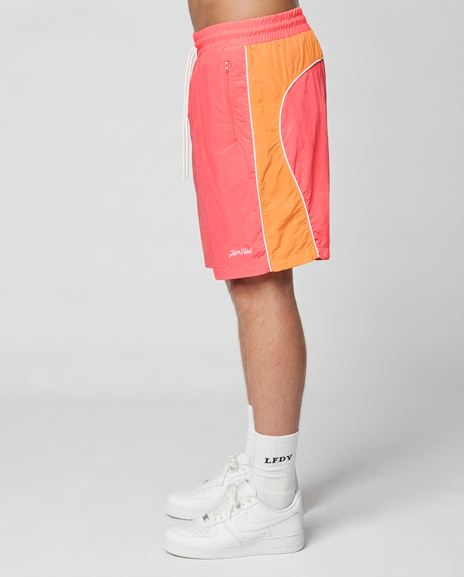 A person stands sideways against a plain light gray background, wearing LIVE FAST DIE YOUNG Beach Shorts in coral and orange with side pockets and elastic waistband, white crew socks with LPDY text, and white Nike sneakers.