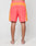 A person stands barefoot, facing away, wearing LIVE FAST DIE YOUNG Beach Shorts in bright orange and coral with white piping and a wide elastic waistband, set against a plain light background.