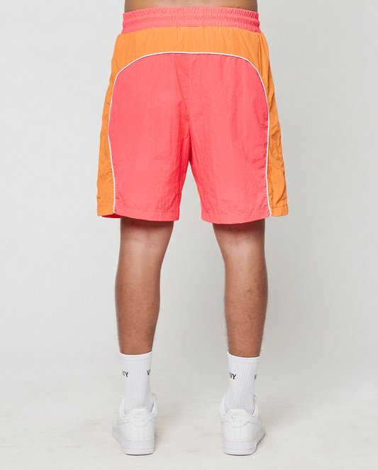 A person faces away, wearing LIVE FAST DIE YOUNG Beach Shorts in orange and pink with Elastikbund and seitliche Eingrifftaschen, paired with white crew socks and sneakers against a plain light background.