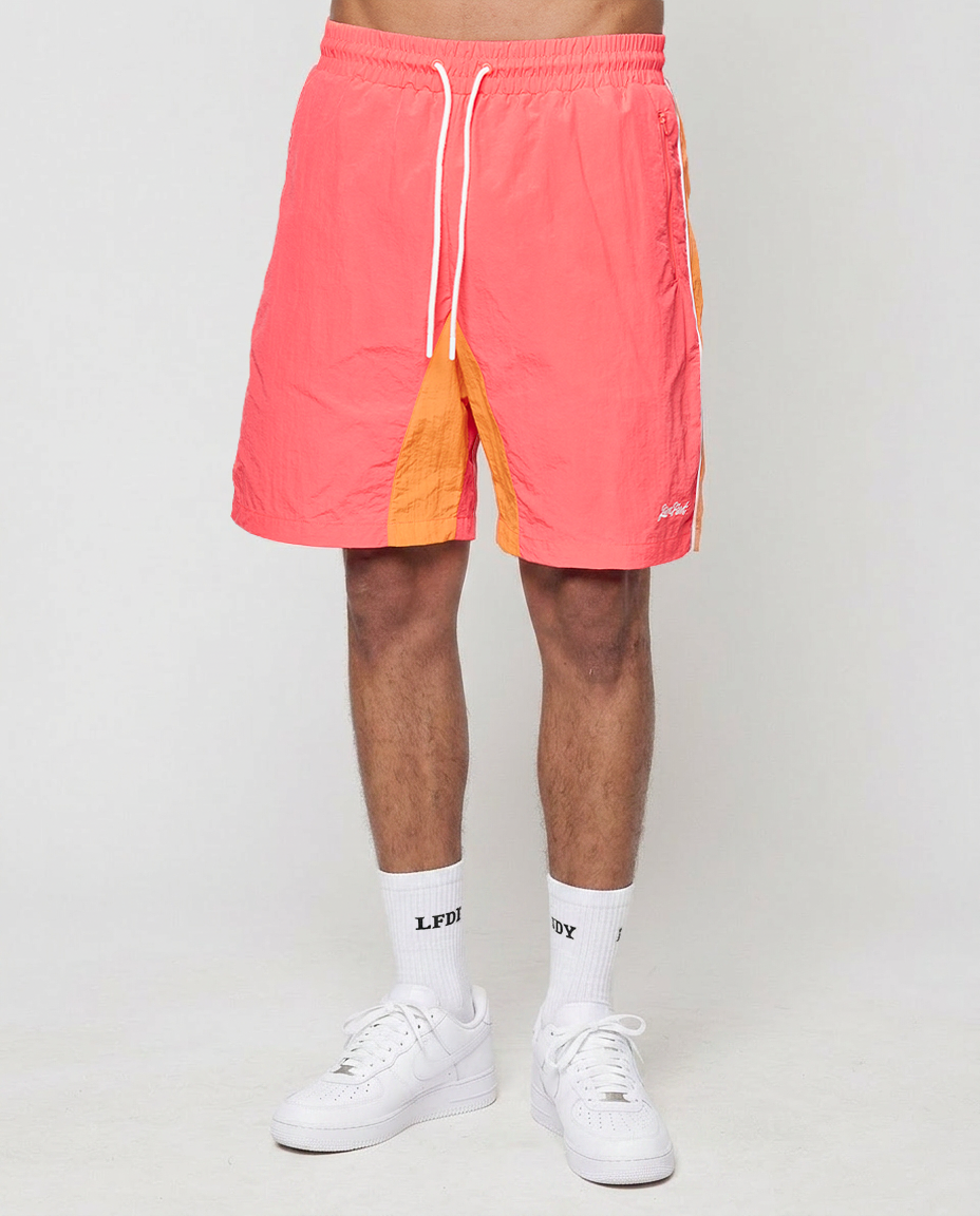 A person in LIVE FAST DIE YOUNG Beach Shorts (Regular Fit, bright coral, elastikbund, seitliche Eingrifftaschen), white LFDY crew socks, and white sneakers stands against a plain light gray background.
