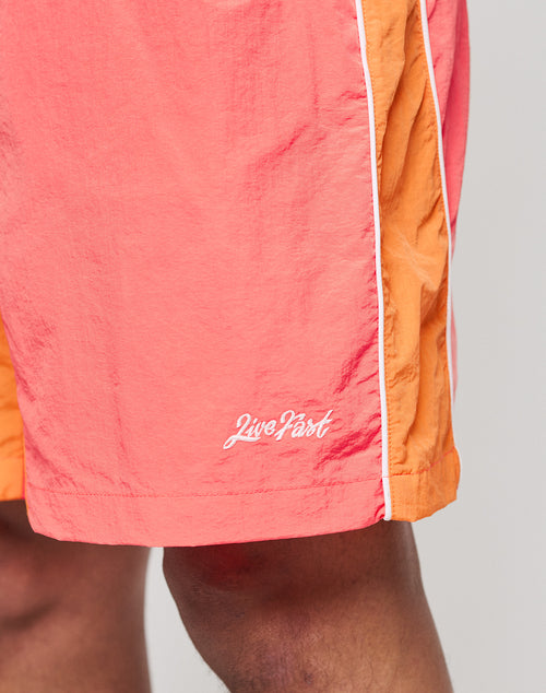 Close-up of a person wearing LIVE FAST DIE YOUNG Beach Shorts in orange and coral with white piping. Features a wide elastic waistband, zippered side pockets, and Live Fast embroidered in white on the left leg.