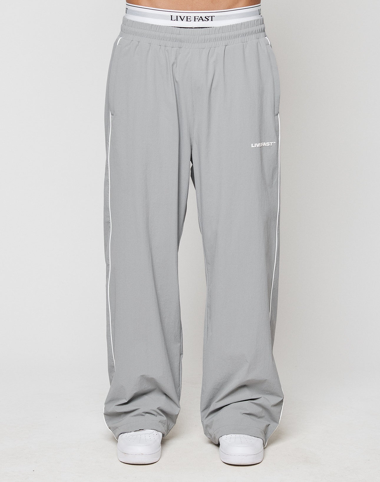 A person wears light gray LIVE FAST DIE YOUNG LFDY Ripstop Trackpants with white side piping, white sneakers, and LIVE FAST silikonprint logos on the waistband and left thigh, set against a plain light background.