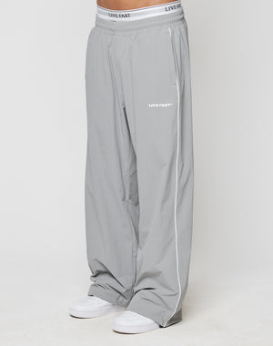 A person wears wide fit, light gray LFDY Ripstop Trackpants by LIVE FAST DIE YOUNG with Elastik-Bund and LIVE FAST silikonprint on waistband and thigh, paired with white sneakers against a plain white background.