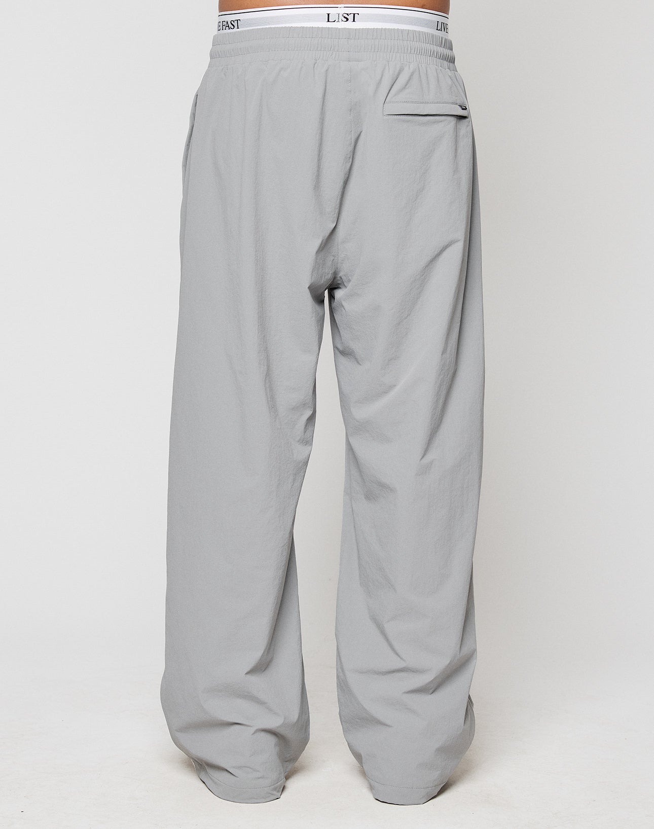 A rear view of a person wearing LIVE FAST DIE YOUNG LFDY Ripstop Trackpants in light gray, featuring an elastic waistband, single back pocket, and wide fit silhouette against a plain white background.