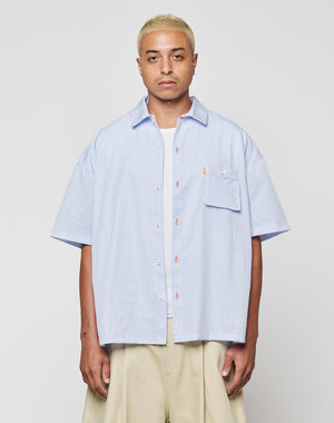 A person with short blonde hair wears the LFDY Striped Boxy Shirt by LIVE FAST DIE YOUNG in light blue and white stripes with a chest pocket and subtle logo, paired with beige wide-leg pants, against a plain light gray background.