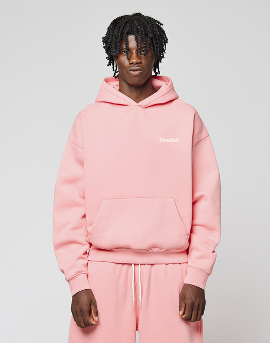 A person with short, curly hair wears an oversize fit light pink Basic 360 Hooded sweatshirt and matching sweatpants from LIVE FAST DIE YOUNG, featuring subtle topstitching and “LiveFast” in small white letters on the chest. The background is plain.