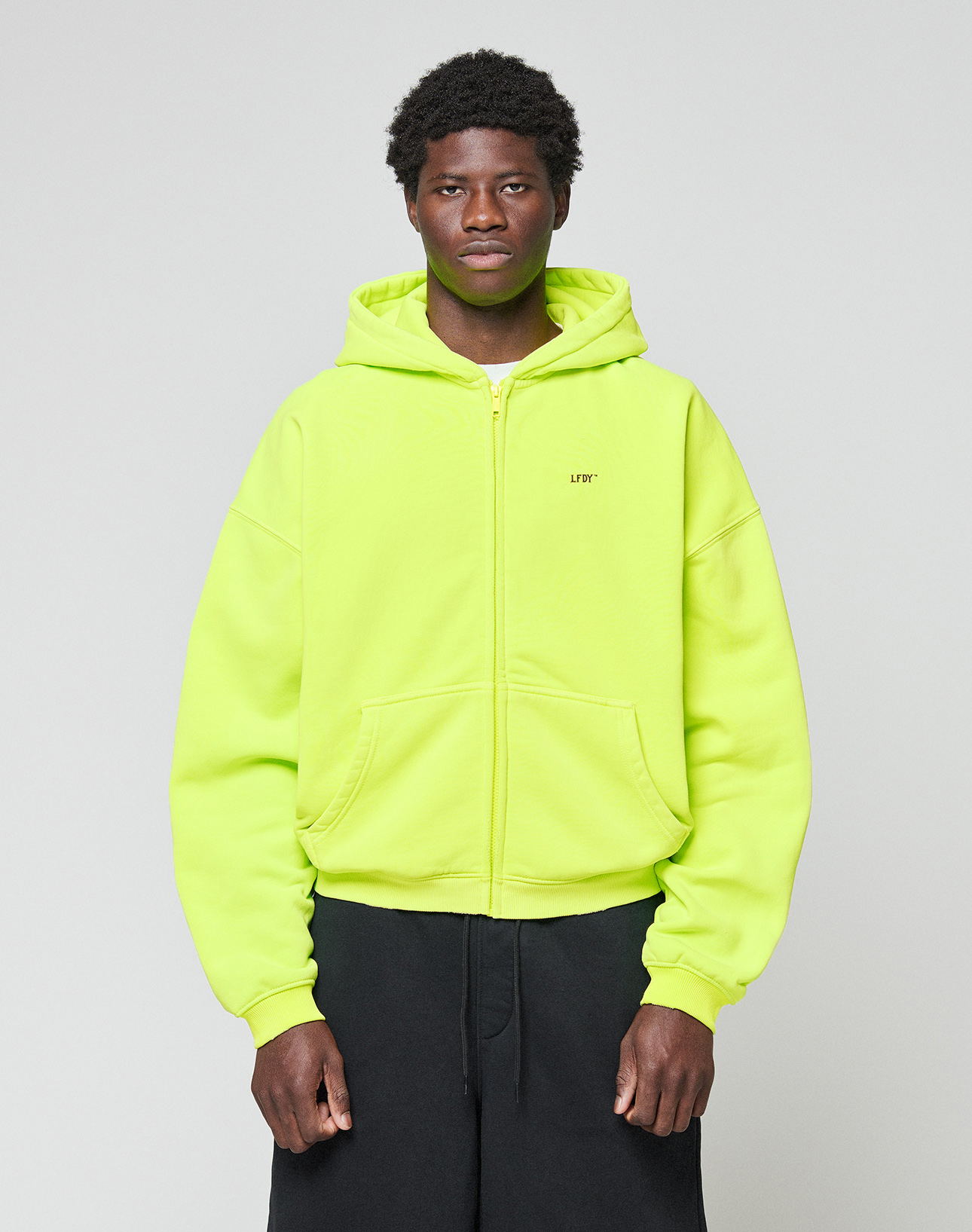 A person stands against a plain light gray background wearing the LIVE FAST DIE YOUNG Basic Hooded Zip in bright neon yellow with black pants. The zip-up hoodie features an oversized fit, and they look straight at the camera with a neutral expression.