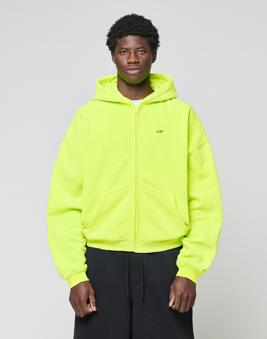 A person stands against a plain light gray background wearing the LIVE FAST DIE YOUNG Basic Hooded Zip in bright neon yellow with black pants. The zip-up hoodie features an oversized fit, and they look straight at the camera with a neutral expression.