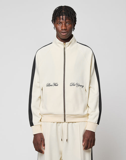A man wears the cream-colored LFDY Striped Track Jacket by LIVE FAST DIE YOUNG with black raglan stripes and matching pants. "Live Fast and Die Young" is embroidered on the chest. He stands against a light gray background.