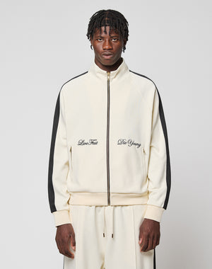 A man wears the cream-colored LFDY Striped Track Jacket by LIVE FAST DIE YOUNG with black raglan stripes and matching pants. "Live Fast and Die Young" is embroidered on the chest. He stands against a light gray background.