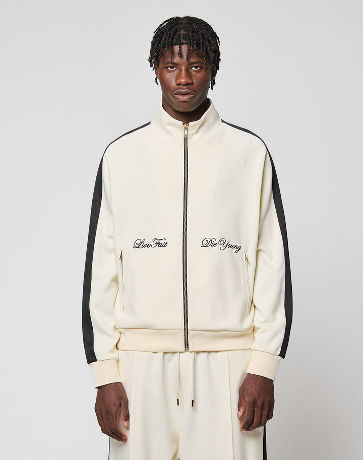 A man wears the cream-colored LFDY Striped Track Jacket by LIVE FAST DIE YOUNG with black raglan stripes and matching pants. "Live Fast and Die Young" is embroidered on the chest. He stands against a light gray background.