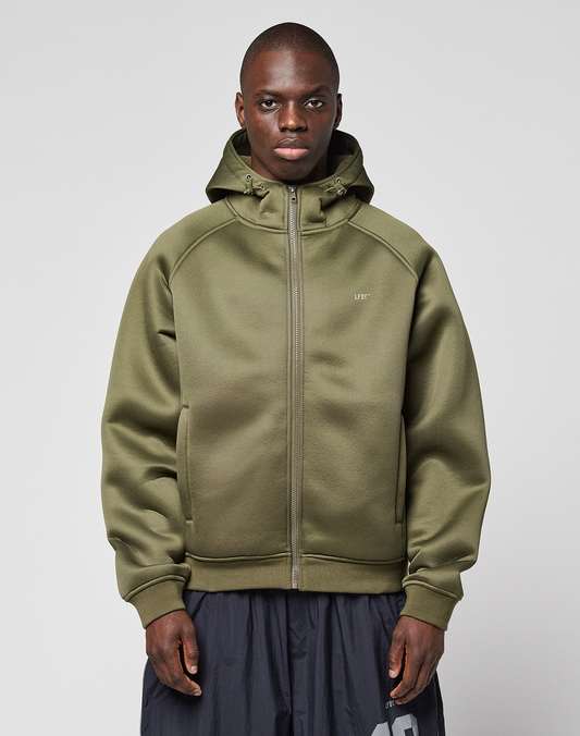 A person stands facing forward, wearing the LFDY Scuba Hooded Zip by LIVE FAST DIE YOUNG in olive green with a hood and zip pockets, regular fit, paired with dark gray pants against a plain light gray background.