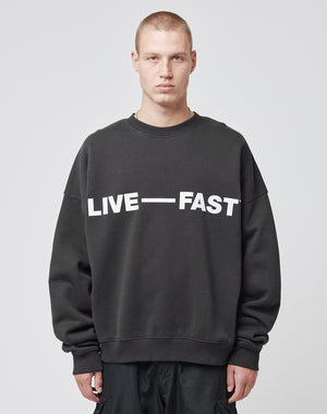 A person with short blond hair wears the LIVE FAST DIE YOUNG Live – Fast Sweater, an oversize fit black sweatshirt with LIVE — FAST in bold white letters, standing against a plain light gray background.